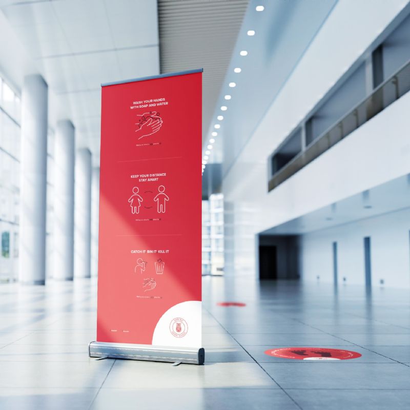 Red roller banner in an atrium with a red footprint floor sticker next to it. Banner depicts 3 COVID-19 awareness messages. 